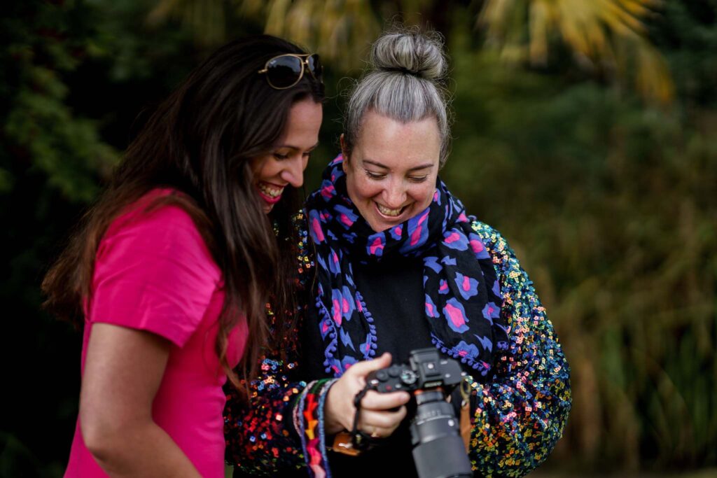 A white female photographer with grey hair and a sparkly jumper shows her camera to her client, a white woman in a pink dress with dark brown hair.