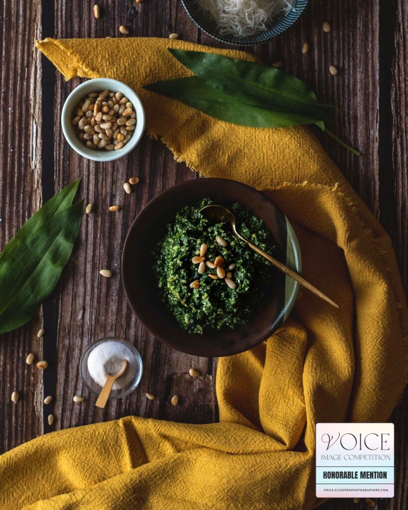 Food photography flatlay of a wooden table full of ingredients and a bowl of green wild garlic pesto, draped with a yellow cloth, taken from above.