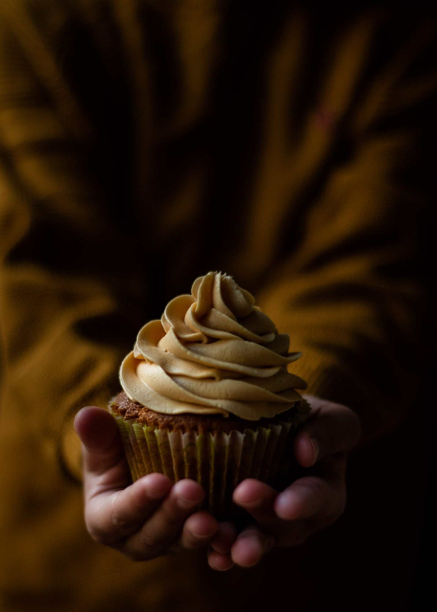 A caramel coloured salted caramel cupcake being held by a child's hands wearing a caramel coloured jumper.