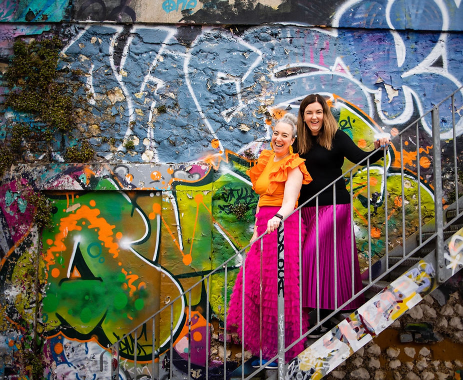 Two female photographers in vibrant clothing, posing in front of colourful graffiti in Brighton